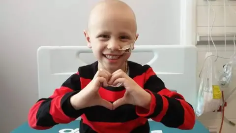 A smiling Ben Crowther wears a red and black striped jumper. He is sitting on a hospital bed with an oxygen tube connected to his nose. He is smiling and making a heart shape with his hands. 