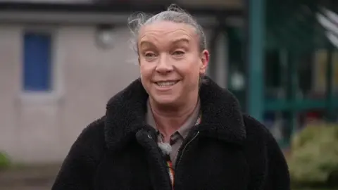 Tamsyn Anderson, chief operating officer for Cornwall Partnership NHS Foundation Trust. She is wearing a large black fleece coat and has her grey hair pulled back into a ponytail. She is stood in front of a hospital building.