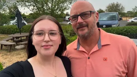 A selfie of a young woman with long dark hair, a black top and light brown glasses, and a bald man with black glasses and grey facial hair wearing a salmon pink t-shirt