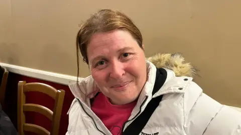 John Devine/BBC Mary Donger, 40, has sandy coloured hair parted in the middle of her head, she is wearing a white padded coat with a fur hood, with a red top underneath. She has a magnolia and burgundy wall behind her.