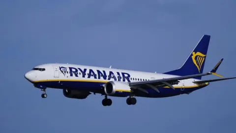 PA Media A Ryanair flight seen flying against a blue sky with the gold harp-style branding as its livery on the back of the plane and Ryanair written on its side 