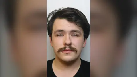 Close up image of young man with dark hair and bushy moustache wearing black t-shirt looking to camera
