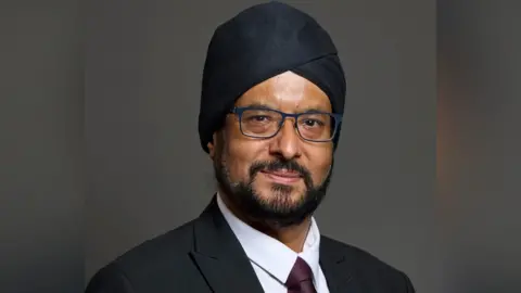 UK Parliament Warinder Juss has a short black beard and moustache and blue glasses. He is looking at the camera with a slight closed mouth smile. He is wearing a black turban, black blazer, white shirt and brown tie.