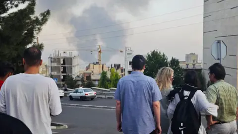 Getty Images People stand with their backs to the camera as they look at smoke rising in the horizon in Tehran on Monday