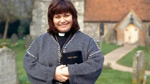 Actress Dawn French, who played Vicar Geraldine Granger in the BBC sitcom The Vicar of Dibley, stands outside St Mary the Virgin Church in Turville. She is holding a black leather bible and wearing a golden cross around her neck.