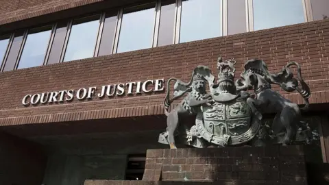 A brown-bricked building with a statue of a coat of arms on a plinth outside.
