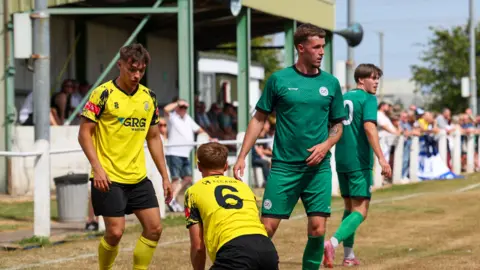 Button Oak Photography Two players wearing green shirts and two in yellow shirts are on a pitch. In the blurred background some people are in a covered area watching the game, while others are standing next to the pitch. 