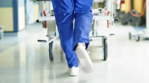 In a generic image of a hospital, a member of nursing staff wearing a blue uniform and white trainers walks down a corridor while pushing a trolley.
