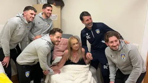 Footballers wearing grey hoodies standing by a blonde woman who is sitting on a sofa with a blanket. They are all smiling at the camera. Two of the men are holding either of her hands while they look into the camera.
