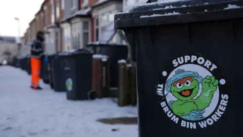 PA Media. Primer plano de un contenedor negro con ruedas y una pegatina de Unite "Apoya a los trabajadores de contenedores de Brum" en el lateral. Al fondo se ve una hilera de casas adosadas y un hombre recogiendo un contenedor negro.