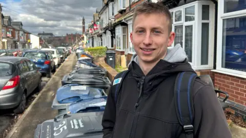 A man, who is wearing a black jacket and carrying a rucksack, is standing in front of dozens of bins in a street lined with terraced houses and cars.