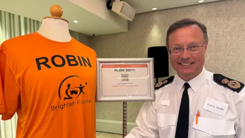 Chief Officer Robin Smith stands next to a mannequin wearing an orange “Brighter Futures – ROBIN” T‑shirt, with a framed sign reading “Robin Smith” displayed beside them.