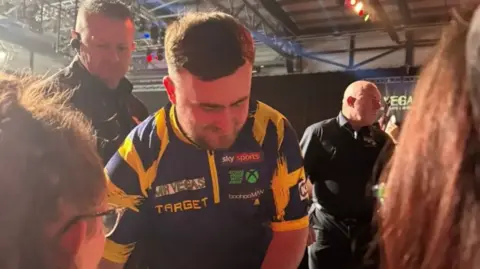 Luke Littler in his signature purple and yellow colours signs fans' autographs in Wolverhampton. Two women with brown hair are facing him and he is flanked by two security guards. 