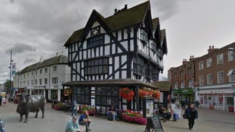 The Black and White House Museum in Hereford. The building has Tudor-style black beams and flowers hanging in baskets around the front. It stands in a pedestrianised square with people sitting on a pedestal in front of it