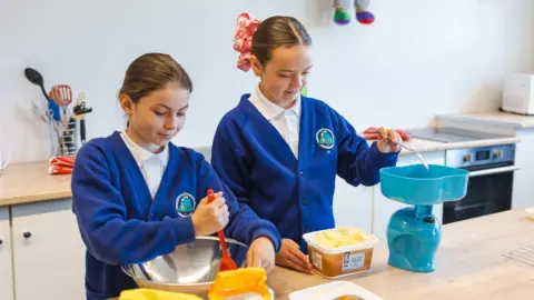 Two girls with brown hair tied back wearing white shirts and blue cardigans. They are in a school kitchen setting with food items and equipment.