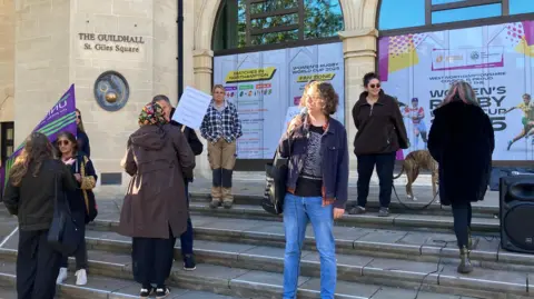 Laura Coffey/BBC A small group of men and women on the steps of Northampton Guildhall. In the middle is a woman in blue jeans and a blue jacket over a black and white jumper with curling light brown hair holding a microphone up to her mouth as she addresses people. 