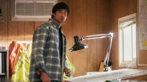 HBO Jacob Elordi as Nate in a checked shirt standing next to a drawing on an architect's desk with an angle-poise lamp in a still from Euphoria