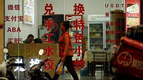 Xiqing Wang / BBC A woman walks down a road in front of the glass walls and doors of a business, with red writing in Cambodian, Chinese and Western scripts. Inside two men appear to work at a wooden desk while fridges with food and drink sit at the back of the store