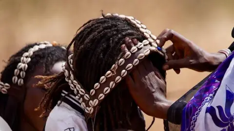 Amensisa IFA / BBC Uma mãe coloca um capacete de concha de cowrie em seu filho para as festividades.