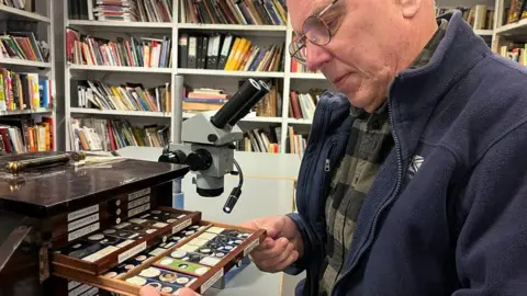 Stuart Robinson/Leeds Museums and Galleries A man with glasses wearing a chequered shirt and blue fleece looks at microscope slides in a small wooden chest