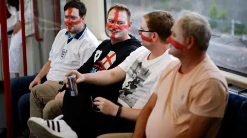 Four men sit together on a train, smiling and talking. Three of them have the red and white flag of England painted onto their faces, and one wears an England football shirt.