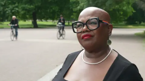 Jennifer Keiza, wearing a black top, large black-framed glasses and a silver necklace, is pictured during an interview in Victoria Park, with grass, trees and a festival site fence behind her.