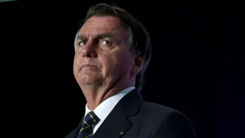 Jair Bolsonaro, who has brown and grey combed over hair and full eyebrows, looks to the side as he stands on a lit stage while wearing a black suit with a white shirt and navy tie with yellow and green stripes