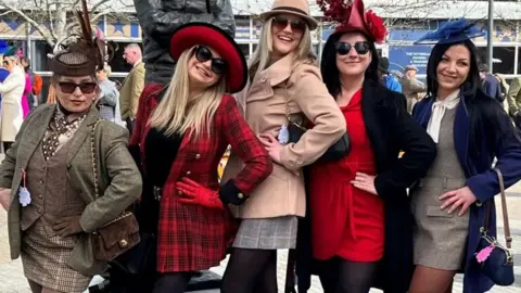 Five women pose flamboyantly at the Cheltenham racecourse. They are all dressed up in tweed and chequered-style minidresses and jackets and wearing hats or fascinators, and sunglasses.