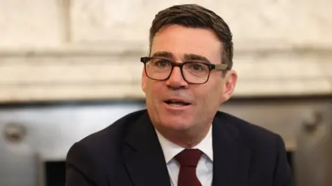 Andy Burnham, a man in a black jacket, with a white shirt and red tie. He has dark hair and is wearing black glasses.