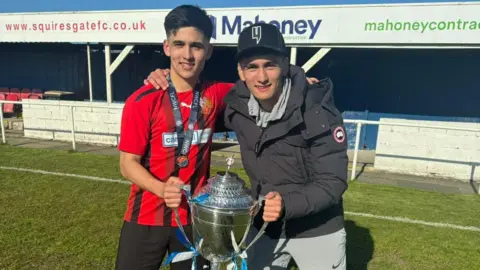 Handout Bryan Ly wears a red and black striped jersey and has black short hair. He is holding a silver trophy with his brother, who wears a black puffa jacket and a black baseball cap. 