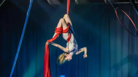 Richard Eyers Photography An aerial performer is on stage, hanging upside-down from a red piece of fabric. Her long blonde hair is in a ponytail.