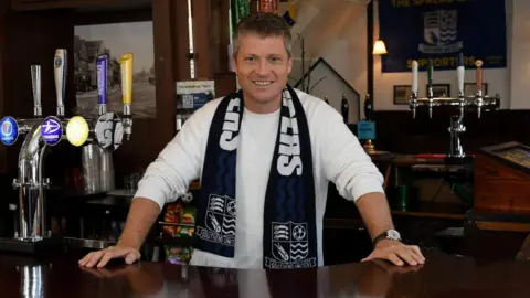 Southend United/Star Pubs Justin Rees, wearing the same outfit as above, standing behind the bar inside the pub.