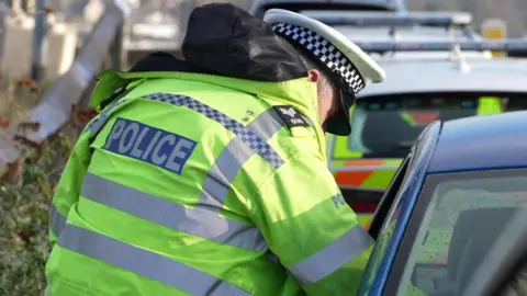 The image shows a roadside scene involving a police officer and a vehicle. The officer is wearing a high-visibility fluorescent yellow jacket with reflective stripes and the word “POLICE” clearly visible on the back. The officer is also wearing a black hood and a white peaked cap with a black-and-white checkered band. The officer is leaning towards the window of a blue car, likely speaking to the driver or checking something inside the vehicle.