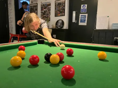 BBC/Amy Holmes A picture of a teenage boy with long blonde hair who is playing pool.