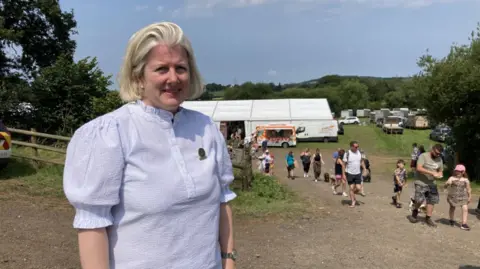 Marcelle Connor in a stiped white and blue button up shirt with frill detail on sleeves and collar looking at the camera with groups of peple in the background and a marquee