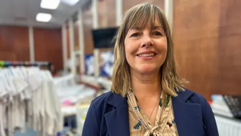 BBC A woman with shoulder length blonde and brown hair is looking at the camera smiling. She is wearing a light brown top and blue blazer. Behind her there is a rail of white shirts and brown wooden walls. 
