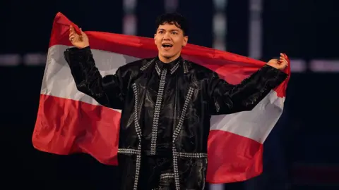 Getty Images JJ representing Austria walks on stage during the Grand Final of the 69th Eurovision Song Contest Opening Ceremony at St. Jakobshalle on May 17, 2025 in Basel, Switzerland. He has dark hair and wearing dark clothing. He he is holding the Austrian flag.