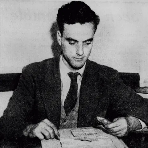 University of Sydney Archives An old black and white photo of Prof Macmillan in a suit, shirt and tie, looking at documents while sitting at a desk