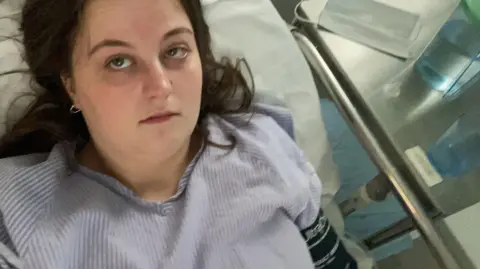 Ella Moulsdale Image of a girl with long brown hair, wearing a blue and white stripe hospital gown who is lying in a hospital bed.