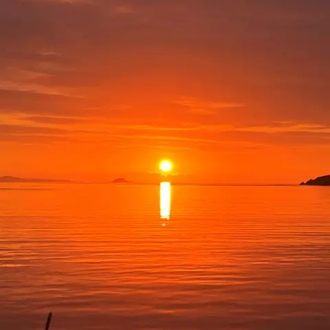 Robert Thomason A sunset over calm water, with the sun low on the horizon and its reflection stretching across the sea. The sky is filled with orange and red tones, and small silhouettes of land are visible in the distance.