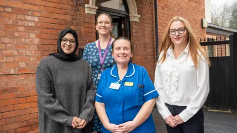 City of Wolverhampton Council Four women are standing outside a brick building, containing a door, in front of a black gate and looking at the camera. They are wearing glasses apart from the woman third from the left, who is in a blue nurse's uniform. The woman far left is in grey, the next person is wearing blue and the woman on the right is in white. 