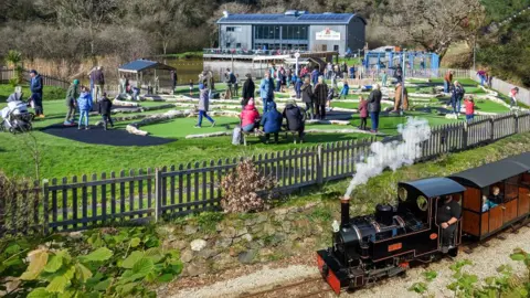 Lappa Valley, with many people enjoying various activities. A miniature steam train with a black engine and wooden passenger cars is on a narrow-gauge railway track in the foreground, emitting white smoke. A wooden fence runs parallel to the track. In the middle is a mini-golf course. In the background, a modern building with a blue roof and large windows overlooks a pond, with an outdoor seating area. There is also a children's playground with blue equipment on the right. The surroundings also feature green trees, bushes, and a natural landscape.