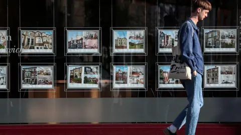 A man wearing blue jeans and a blue shirt walks past an estate agent window displaying home listing photos.