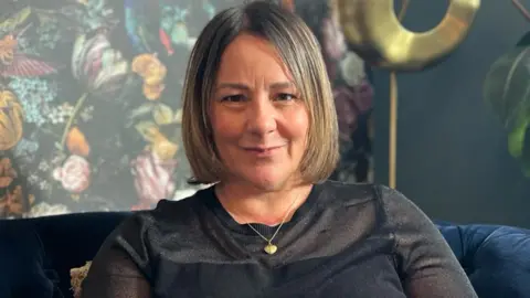 A head shot of Sue Greenwood. She has mid-length blonde/brown hair and is sitting on a sofa in front of green floral wallpaper. She is wearing a long sleeve black top and a gold necklace. 
