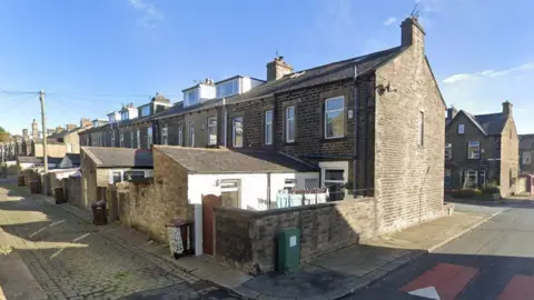 Alley next to row of terraced houses and backyards in Colne. The houses are a dark brown brick, and the alleyway is cobbled. Most of the houses on the row have a loft extension and small backyards. One has a washing line with washing pegged up in the sunshine.