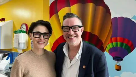 Emma Willis pictured smiling. She is wearing thick framed black glasses and a brown jumper. Ed James is also smiling and wears black frame glasses, a white shirt and blue blazer. They stand in front of a hot air balloon painted wall