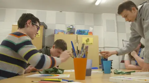 Young people around a table filled with paintbrushes, cups and pictures.