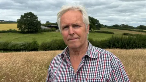 There is a man with light white hair, brushed back. He has a light stubbly beard. He has a blue, red and grey chequered t-shirt, with white squares in the middle. He has two buttons undone. Behind him is yellow grazing land, a dark green hedgerow and fields behind him.