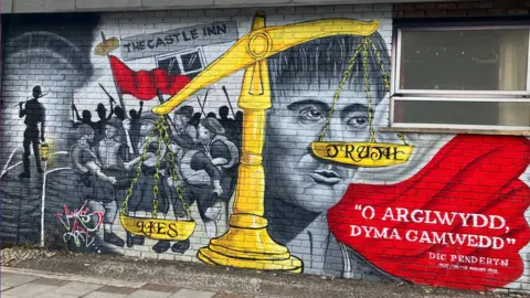 Brick wall with black and white graffiti depicting the face of Dic Penderyn, the scene of the Merthyr Rising with a number of people holding sticks in the air and a red flag. There's also a large golden scales in the centre of the mural with the words Lies and Truth written each side. On the lower right side of the mural is a block of red with the words "O Arglwydd dyma gamwedd" ("Oh Lord this is iniquity") which are believed to be the final words uttered by Dic Penderyn before he was executed.