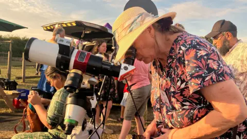 Kit Taylor/BBC A woman in a floral top peers through a telescope